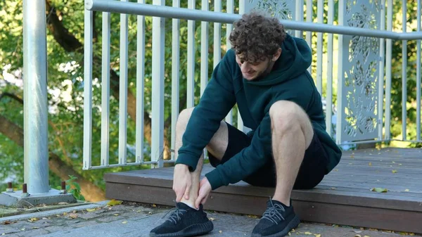 Young man sitting on stairs, feeling pain in his leg. Closeup feet, arms, ankles, fingers. Guy massaging his feet. Sportsman in black spot shorts, hoodie, training shoes relaxing and massaging leg.