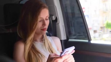 Takside seyahat eden genç bir kadın arabanın arka koltuğunda oturuyor, cep telefonunda daktilo yazıyor. Yaşam tarzı, yolculuk, teknoloji konsepti. Gerçek zamanlı.