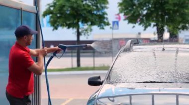 Üniformalı bir erkek kurye arabasını açık hava yıkama istasyonunda su tabancasıyla yıkıyor. self servis, araba yıkama konsepti. Gerçek zamanlı.