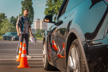 Sürücü kursunda bir bayanın nasıl park ettiğine bakarken elinde bir pano vardı. Arabanın yanında trafik konileri var. Ehliyet sınavı, ehliyet kursu, sınav konsepti.