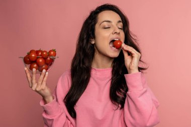 Kameranın önünde pembe arka planda kırmızı domates ısıran esmer bir kadının belden yukarı portresi. Çiğ yiyecek ve sağlıklı beslenme konsepti