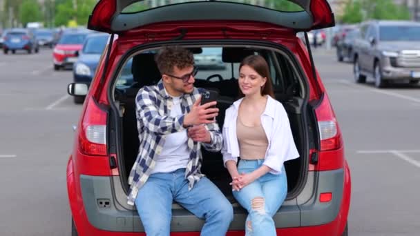 Усміхнена Молода Пара Сидить Багажнику Парковці Людина Тримає Мобільний Телефон — стокове відео