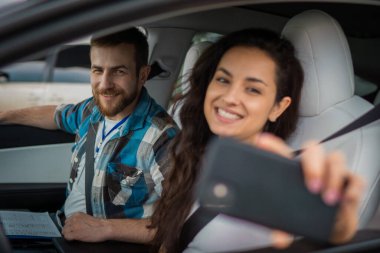 Gülümseyen, selfie çeken ve cep telefonu tutan genç bir bayan, arabada yanında oturan erkek eğitmen, form tutuyor. Ehliyet sınavı, ehliyet kursu, sınav konsepti.