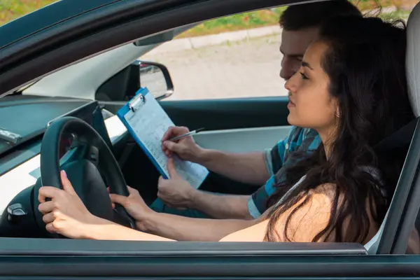 Happy young woman learning to drive a car while driving school instructor holding clipboard. Driving test, driver courses, exam concept