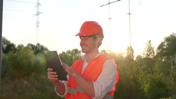 デジタルタブレットで作業しながら屋外に立っている笑顔の男性電気エンジニア 高圧パワーライン エネルギー産業コンセプト スローモーション — ストック動画