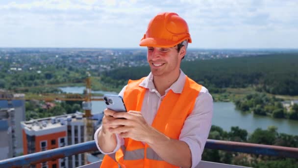 携帯電話にタイピングしながら建設現場の屋根に立っているハッピーな若いビルダー ビジネス ビルディング 産業コンセプト スローモーション — ストック動画