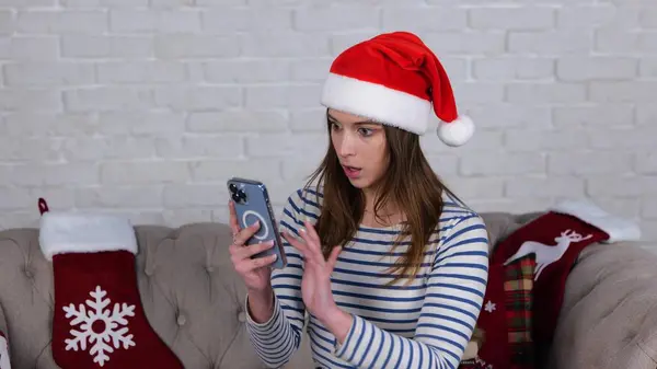 Surprised lady in Christmas hat using smartphone, reading message while sitting on couch at home. Woman looking at phone screen and reading shocking news. Slow motion video