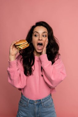 Korkmuş bir kadın pembe arka planda dururken elindeki çizburgere bakıyor. Fast food konsepti