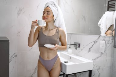 İç çamaşırlı, gözleri kapalı elinde fincan ve çay tabağı tutan ve banyoda aynası olan büyük lavabonun yanında duran genç bir bayan. Yaşam tarzı kavramı