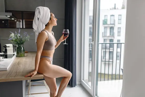 Back view of woman in underwear leaning on the table in the kitchen at home and holding glass of wine. Lady looking out the window