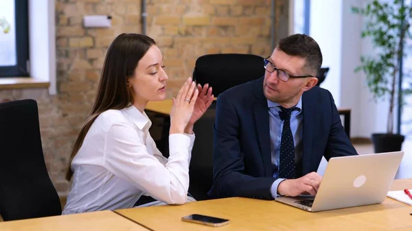 Caucasian man and woman are sitting in modern office on table. Businesswoman in white shirt explaining her ideas to man colleague. Man in blue shirt and black suit looks happy, he is smiling.