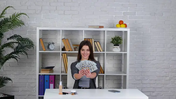 Smiling woman showing fan from dollars in hands and throwing dollar bills in the air while sitting at the desk and looking at camera. Slow motion video