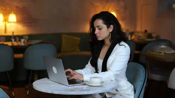 Pretty young lady typing on laptop while having coffee break in restaurant, drinking cappuccino. Copy space. Business, lifestyle concept. Slow motion