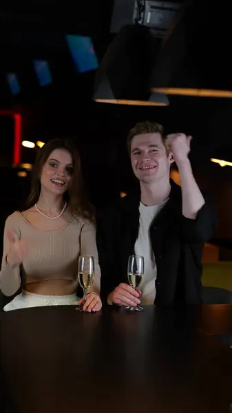 Smiling guy and lady with glasses of champagne and showing thumbs up, sitting and posing in the nightclub. Lifestyle, party, vertical, social networks concept. Slow motion