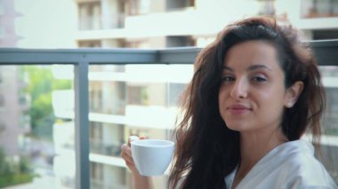 Elinde çay fincanı tutarken gülümseyen neşeli, genç ve çekici bir bayan. Güzel, beyaz, mutlu bir kadın apartmanın balkonunda kahve içiyor. Kamera geri gidiyor. Yavaş çekim