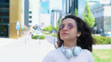 Güneş gözlüklü, neşeli, genç bir kadın şehirde yürüyor ve etrafa bakıyor. Beyaz, mutlu bayan öğrenci sokakta yürüyor. Yakın çekim. Güneşli bir yaz günü. Açık hava konsepti. Yavaş çekim