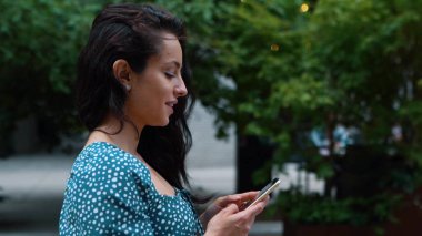 Young beautiful attractive woman texting message on smartphone on social media while walking in town outdoors. Close up. Side view. Pretty happy brunette female browsing online on cellphone outdoor