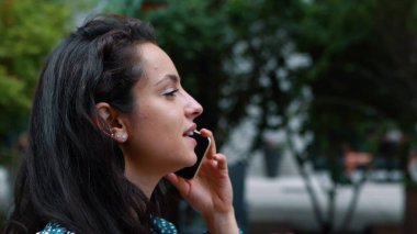 Side view of young attractive female smiling and speaking on mobile phone while walking in street. Close up of beautiful positive woman calling on cellphone outside in good mood. Communication concept