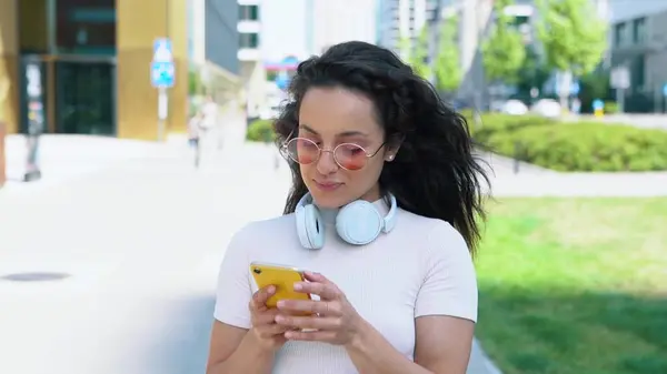 Beautiful casual Caucasian woman in pink glasses walking in the street in city and typing on mobile phone. Close up. Cheerful young dark-haired female student texting on smartphone. Urban concept