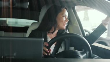 Gülümseyen genç iş kadını arabanın camından bakıp el sallıyor, şehirde araba sürüyor. Güvenli sürüş, ulaşım, seyahat, iş konsepti. Yavaş çekim