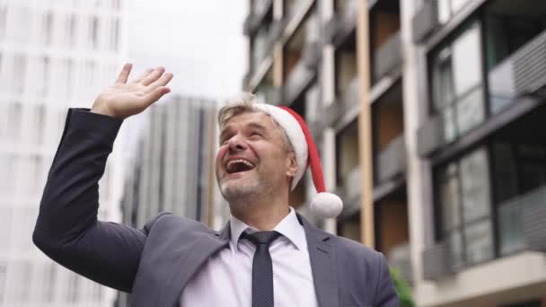 サンタの帽子にスーツを着た笑顔の大人の男は屋外に立って挨拶しながら手を振ります お祝い クリスマス ビジネスコンセプト スローモーション — ストック動画