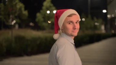 Akşam şehirde yürürken omzunda ceketi olan, Noel Baba şapkalı gülümseyen genç adam. İş, kutlama, yeni yıl konsepti. Yavaş çekim