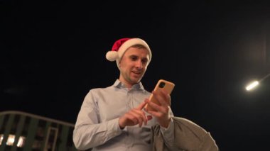 Gülümseyen genç adam dışarıda duruyor ve bayramı kutluyor, birden beşe kadar sayıyor, cep telefonu tutuyor, Noel Baba şapkasını kusuyor. İş, kutlama, yeni yıl konsepti. Yavaş çekim