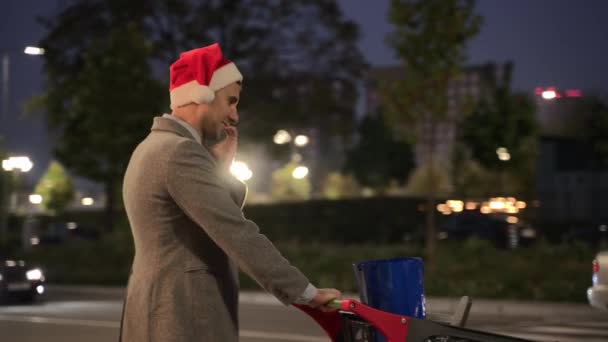 サンタのハッピーな男は スマートフォンで話している間 屋外の彼の車にショッピングカートを押すのが大好きです ビジネス お祝い 新年のコンセプト スローモーション — ストック動画