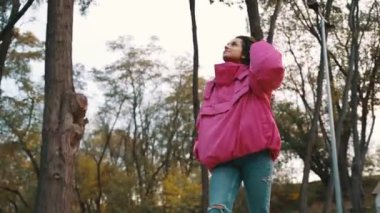 Pembe ceketli gülümseyen şık bayan sonbahar parkında yürüyor ve manzaranın tadını çıkarıyor. Sonbahar sokak modası. Sonbahar sezonu. Gerçek zamanlı.