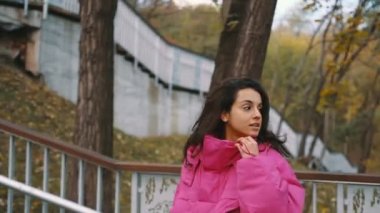 Pembe ceketli genç bayan şehir parkındaki ahşap merdivenlerden çıkıyor. Sonbahar sokak modası. Sonbahar sezonu. Gerçek zamanlı.