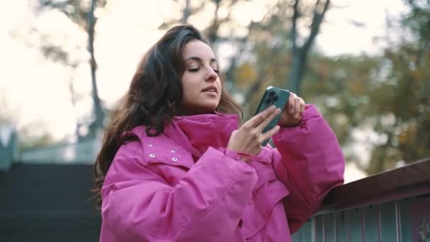 站在城市公园时 穿着粉色夹克的时髦女士在手机上拍照 快乐极了 秋天都市街道时尚风格 秋天的季节实时 — 图库视频影像