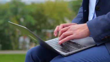 Genç adamın dizüstü bilgisayarda çalıştığı ve klavyeyle parkta oturduğu görüntüleri kapatın. Sokakta bilgisayarda yazan erkek parmakları. İş hayatı. İşçi konsepti. Gerçek zamanlı konsept.