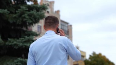 Sokakta yürüyen ve cep telefonuyla konuşan gömlekli bir adamın arka görüntüsü. Şehirdeki bir adam tek başına yürüyor ve cep telefonuyla konuşuyor. İletişim. Telefon görüşmesi konsepti. Gerçek zamanlı konsept.