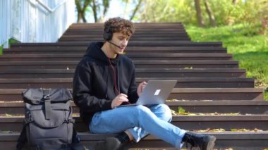 Merdivenlerde oturup, kulaklık takarak internette dizüstü bilgisayarla çalışan mutlu adam. Sonbahar sezonu. Eğitim, çalışma konsepti. Gerçek zamanlı konsept.