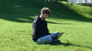 Şehir parkında çimlerde otururken dizüstü bilgisayar ve daktilo kullanan mutlu bir adam. Sonbahar sezonu. Eğitim, çalışma konsepti. Gerçek zamanlı konsept.