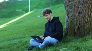 Yazarken dizüstü bilgisayar kullanan gülen adam. Şehir parkındaki ağacın yanında oturan adam. Sonbahar sezonu. Eğitim, çalışma konsepti. Gerçek zamanlı konsept.