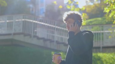 Cep telefonuyla konuşurken ve elinde kahve tutarken şehir parkında yürüyüş yapmaktan zevk alan şık bir adam. Yaşam tarzı. Sonbahar yürüyüşü kavramı. Gerçek zamanlı konsept.