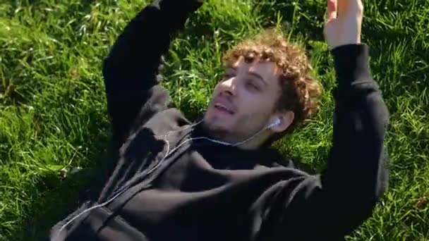 Jovem Sorridente Deitado Gramado Parque Cidade Ouvindo Música Fones Ouvido — Vídeo de Stock