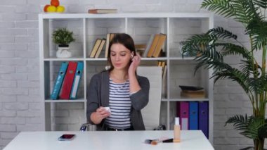 Modern makyaj ve cilt bakım ürünleri hakkında konuşan moda blogcusu evde otururken akıllı telefon ve kablosuz kulaklık kullanıyor.