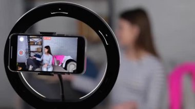 Evde telefon kamerasıyla video kaydederken askılarda şık kıyafetler sergileyen neşeli genç bayan tripoddaki akıllı telefon ekranına odaklanıyor. Gerçek zamanlı video.