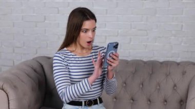 Evdeki bir kanepede otururken akıllı telefondan kötü haberler okuyan Kafkasyalı kadın için endişeleniyorum. Alet kullanırken nahoş haberler okuyan genç bir bayan. Gerçek zamanlı video.