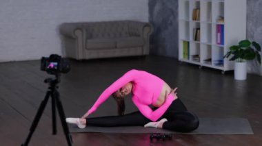 Pembe spor bluz giyen çekici ve odaklanmış bir bayan. Evde bükülüp vlog için spor videoları kaydediyor. Yan kıvrımlar esneme, vücut eğimleri. Gerçek zamanlı video.