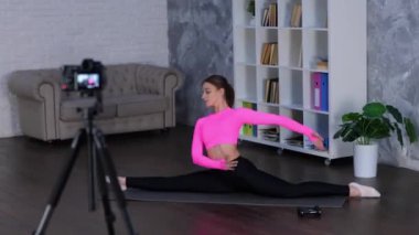 Tozluklu ve üstsüz atletik kadın sicimin üzerinde otururken kadın evde yoga egzersizi yapıyor. Tripodun üzerinde kamera kullanan bir kadın. Spor salonu, spor konsepti. Gerçek zamanlı video.