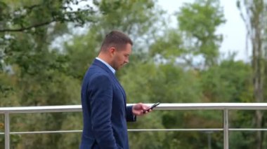 Portre fotoğrafı. Kentteki köprüde takım elbiseli genç beyaz bir adam yürüyor ve akıllı telefondan mesaj atıyor. Gerçek zamanlı konsept. Başarılı erkek girişimci cep telefonuyla dışarı çıkıyor.