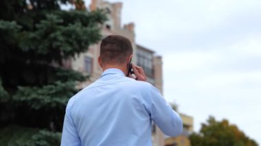 Sokakta yürüyen ve cep telefonuyla konuşan gömlekli bir adamın arka görüntüsü. Şehirdeki bir adam tek başına yürüyor ve cep telefonuyla konuşuyor. İletişim. Telefon görüşmesi konsepti. Gerçek zamanlı konsept.