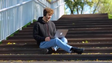 Şehir parkındaki merdivenlerde oturan bir adam dizüstü bilgisayar ve kulaklıkla çalışıyor, video görüşmeleri yapıyor. Sonbahar sezonu. Eğitim, çalışma konsepti. Gerçek zamanlı konsept.
