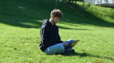 Şehir parkında çimlerde otururken dizüstü bilgisayar ve daktilo kullanan mutlu bir adam. Sonbahar sezonu. Eğitim, çalışma konsepti. Gerçek zamanlı konsept.