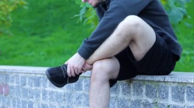 Adam dışarıda ayak bileğini esnetiyor. Green Park 'ta bankta oturan adam bacağına masaj yapıyor. Sporcu şort, kapüşonlu, spor ayakkabılı koşudan sonra ayak ve ayak bileğinde güçlü bir ağrı hissediyor.