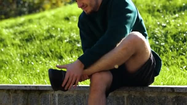 户外活动结束后 运动员坐在绿地公园的长椅上舒展腿 贴身男子穿着短裤 运动鞋 按摩他的腿 感觉他的腿疼痛 真正的 — 图库视频影像