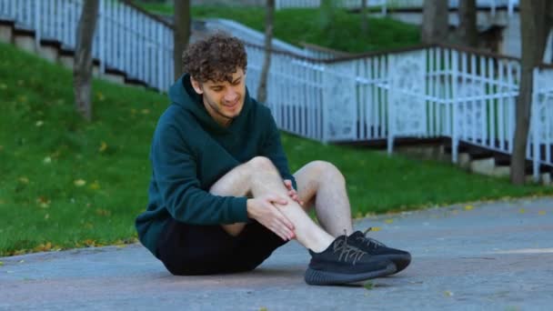 年轻的白人男子坐在地上 伸展他的膝盖 肌肉在绿地公园里 运动员在锻炼前感到腿剧痛 并按摩膝盖和肌肉 — 图库视频影像